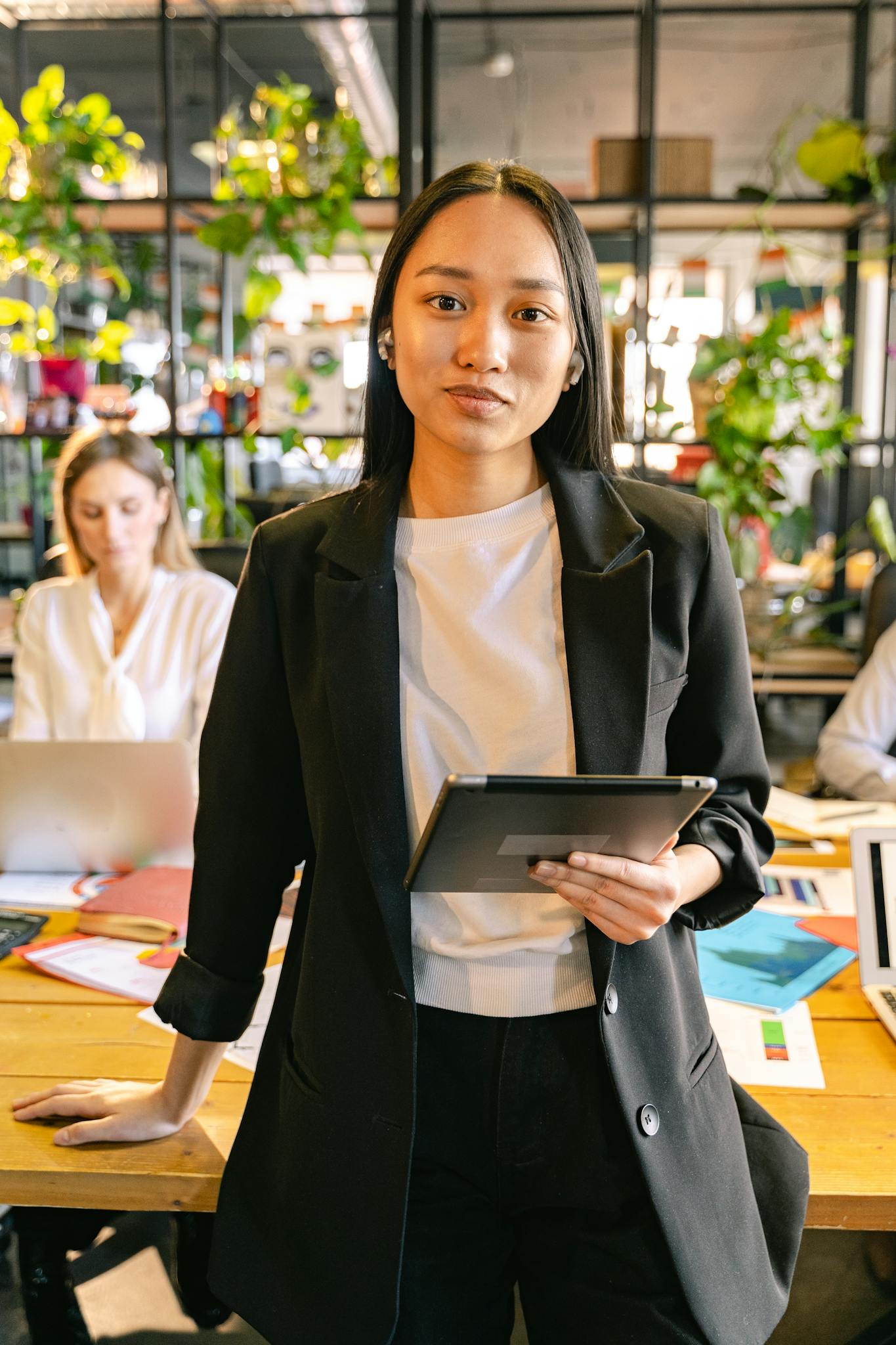 Confident professional woman holding a tablet in a vibrant modern office with colleagues working in the background.